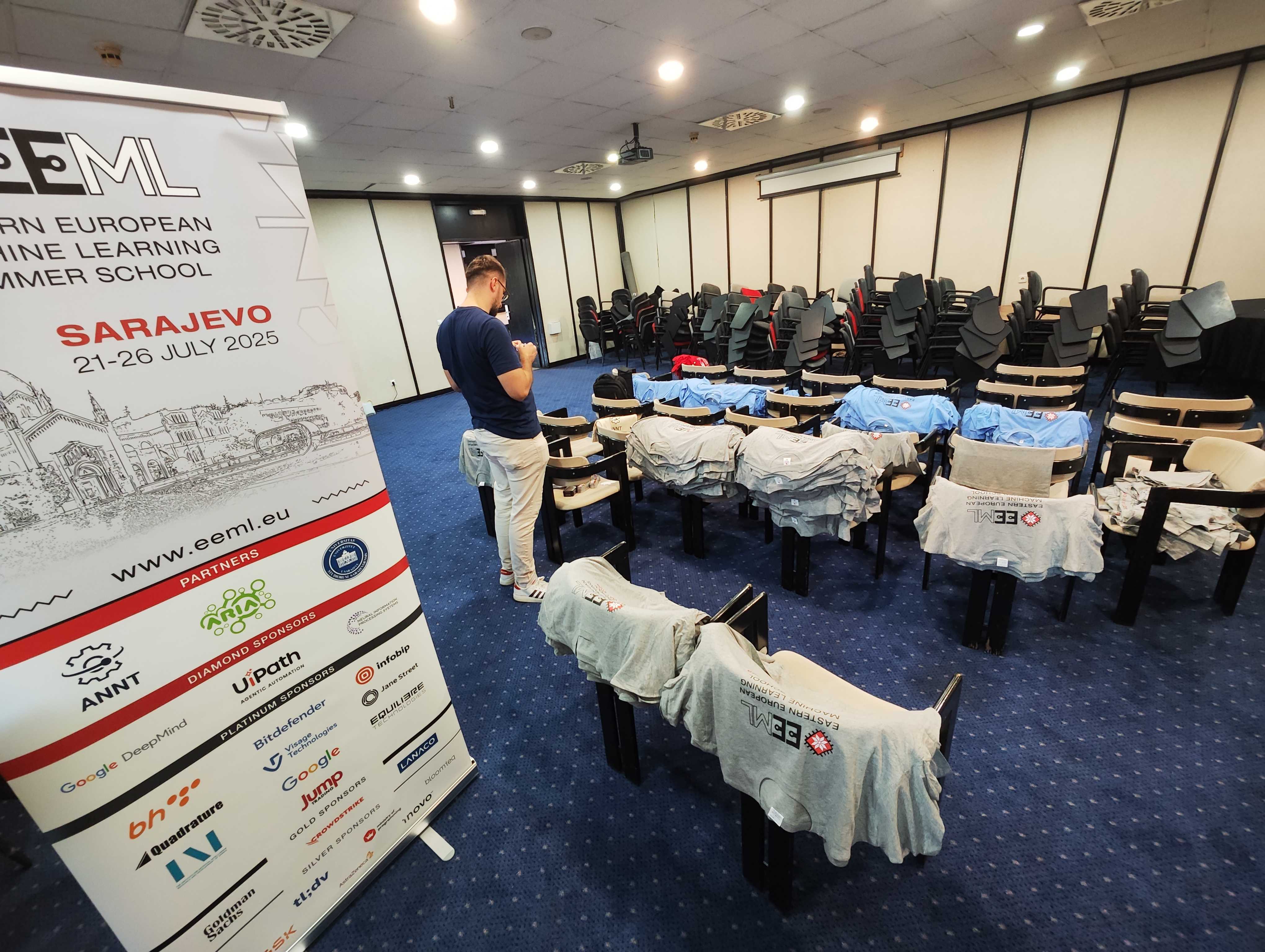 A stack of EEML 2025 T-shirts next to rows of lecture chairs with fold-out tables.